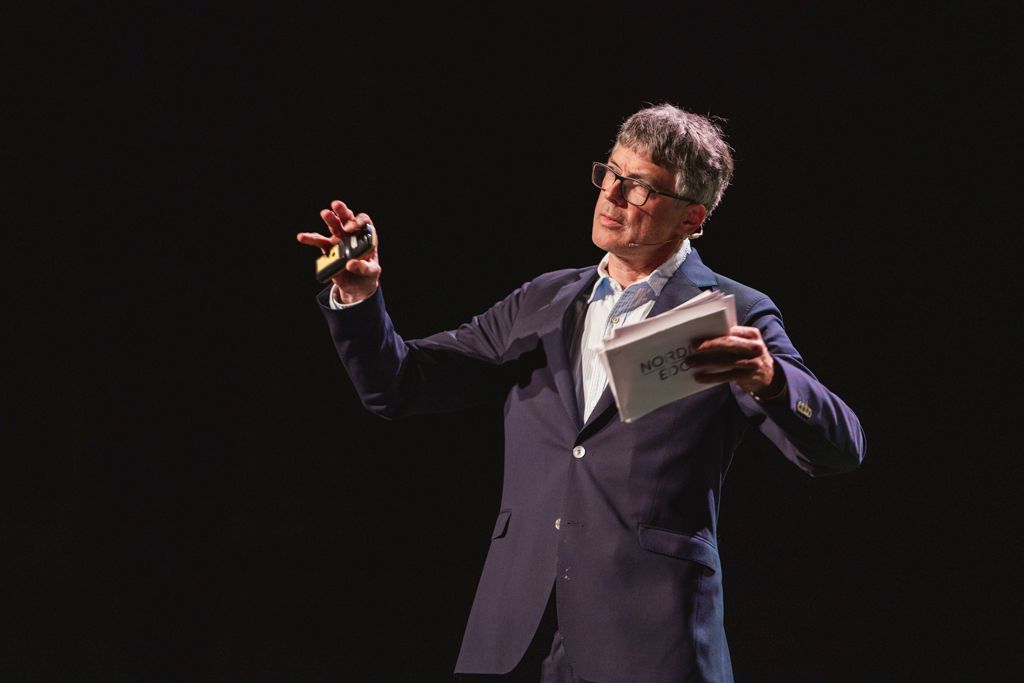 Man in suit gestures, holding a small book, standing on a stage with a dark background.