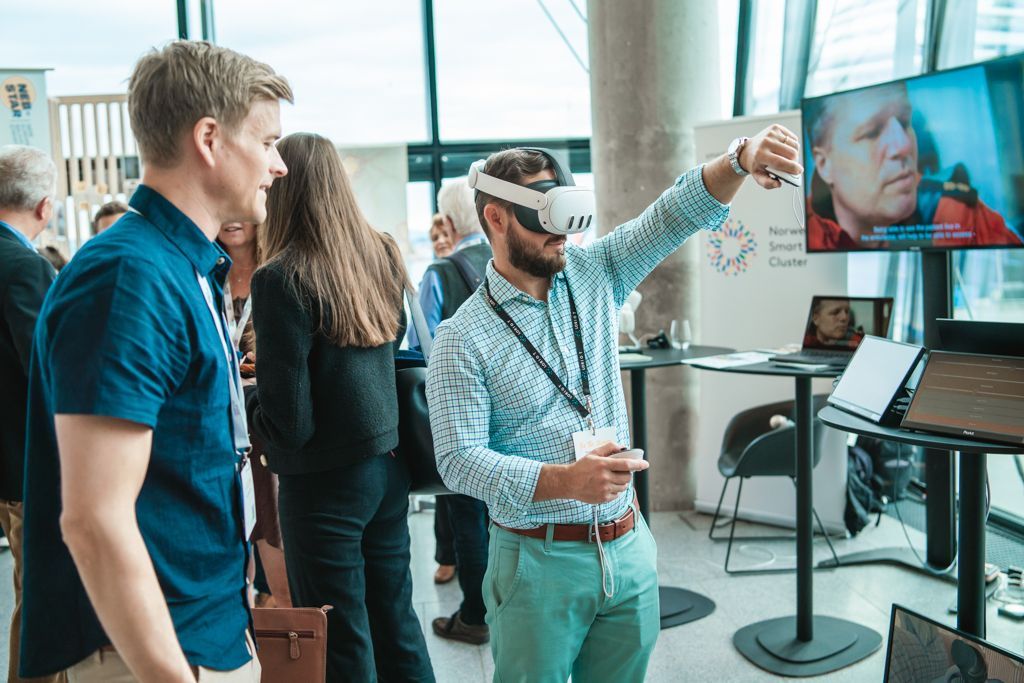 Man in VR headset demonstrates a virtual experience to another man. A monitor displays a person's face.