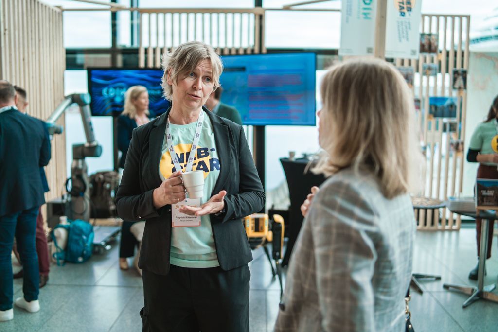 Woman in blazer talking to another woman at an event, holding a mug. Other people and screens in background.