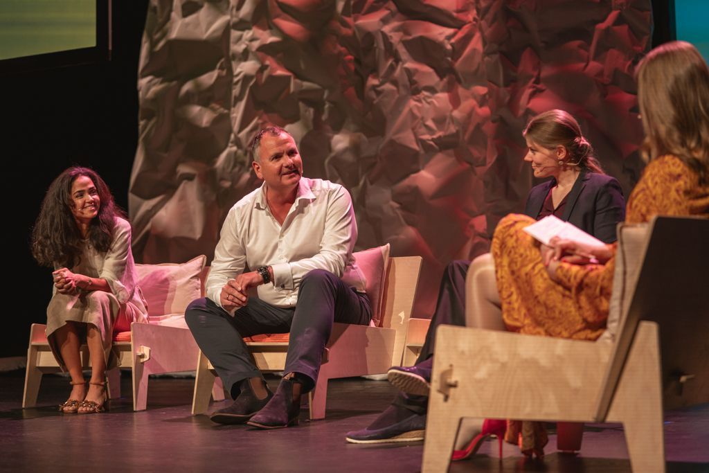Panel of four people on a stage with a crumpled paper backdrop, seated on modern chairs, smiling.