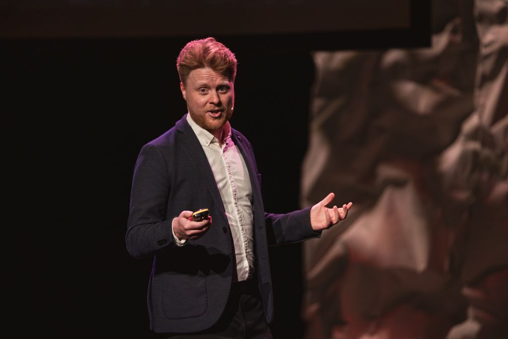 Man in suit giving a presentation, gesturing with hands. Dark background with projection screen.