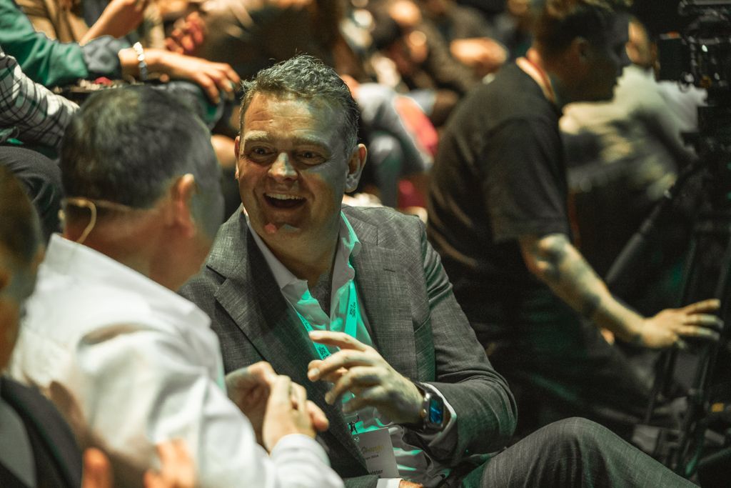 Man in grey blazer laughs while conversing with another man in a white shirt, seated in a crowded room.