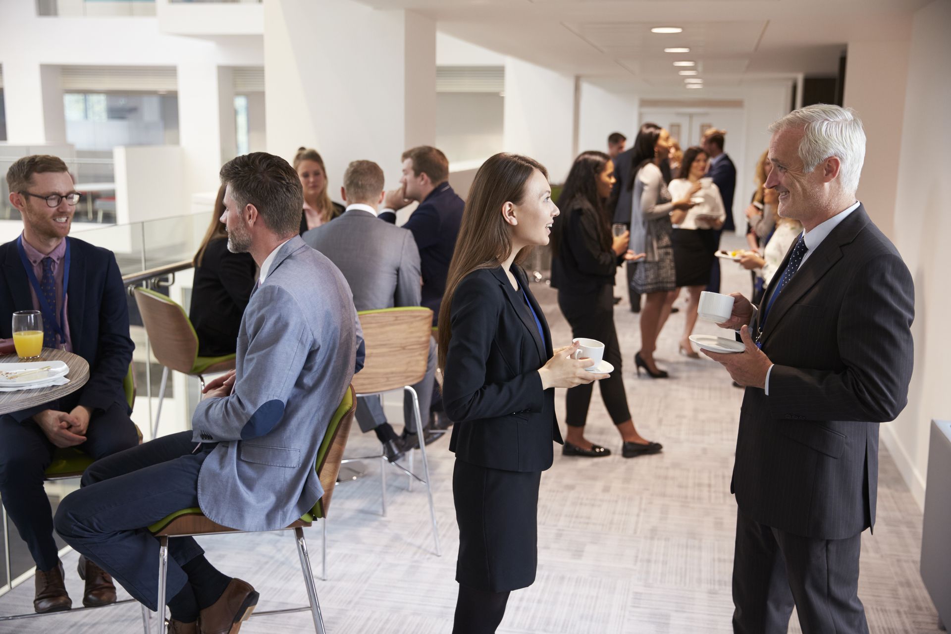 People networking at a business conference, indoors. Several groups chat while holding drinks. Bright, modern setting.