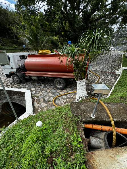 Un camión cisterna rojo está estacionado junto a un canal de carretera y un terraplén cubierto de hierba con tuberías y árboles.