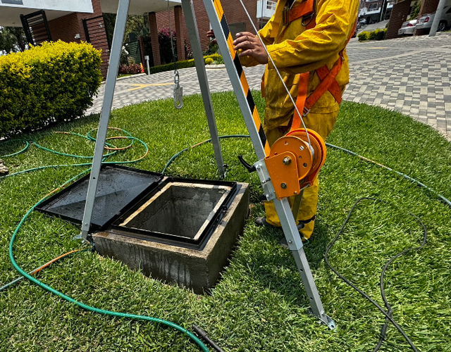 Un trabajador con chaleco naranja baja una tapa de hormigón sobre una alcantarilla en una zona de césped.
