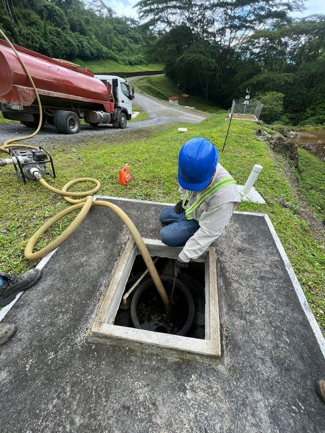 Un trabajador con casco azul inspecciona una alcantarilla abierta al borde de la carretera, junto a un camión de vacío.