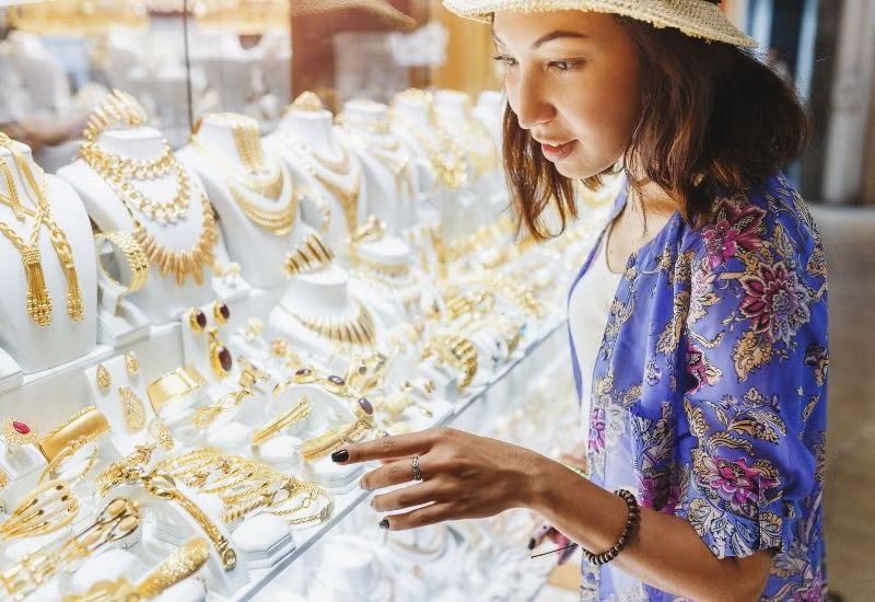 Woman browsing gold jewelry in a brightly lit store display, wearing a blue patterned top and straw hat
