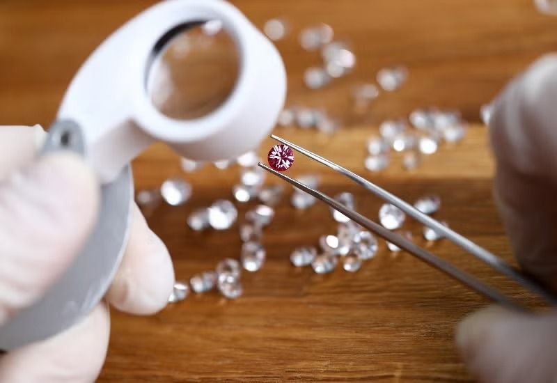 Gloved hands use tweezers and a magnifier to inspect a small gemstone on a wooden table.
