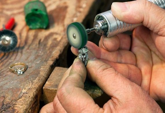 Hands use a rotary tool to polish a small piece of jewelry on a wooden workbench.