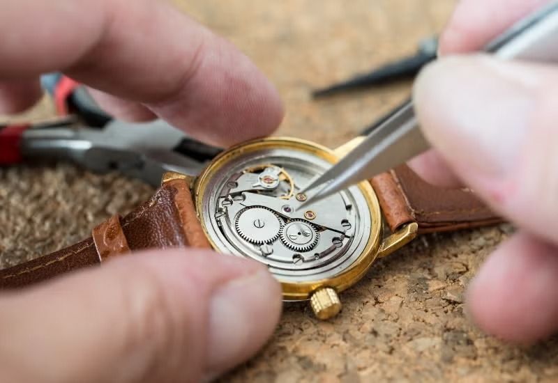 Hands using tweezers to repair a gold wristwatch on a workbench
