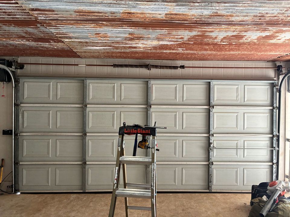 Garage with a gray paneled door, rusted ceiling, and a step ladder centered in front.