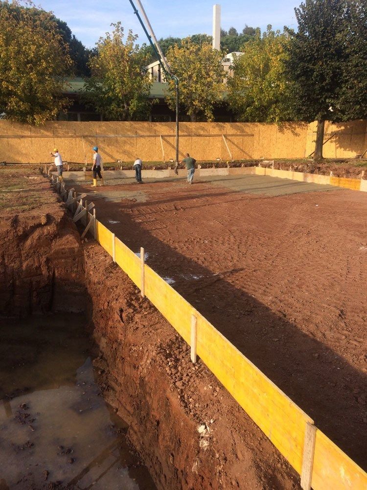 Construction site with workers leveling a large concrete slab beside an excavated pit and yellow formwork