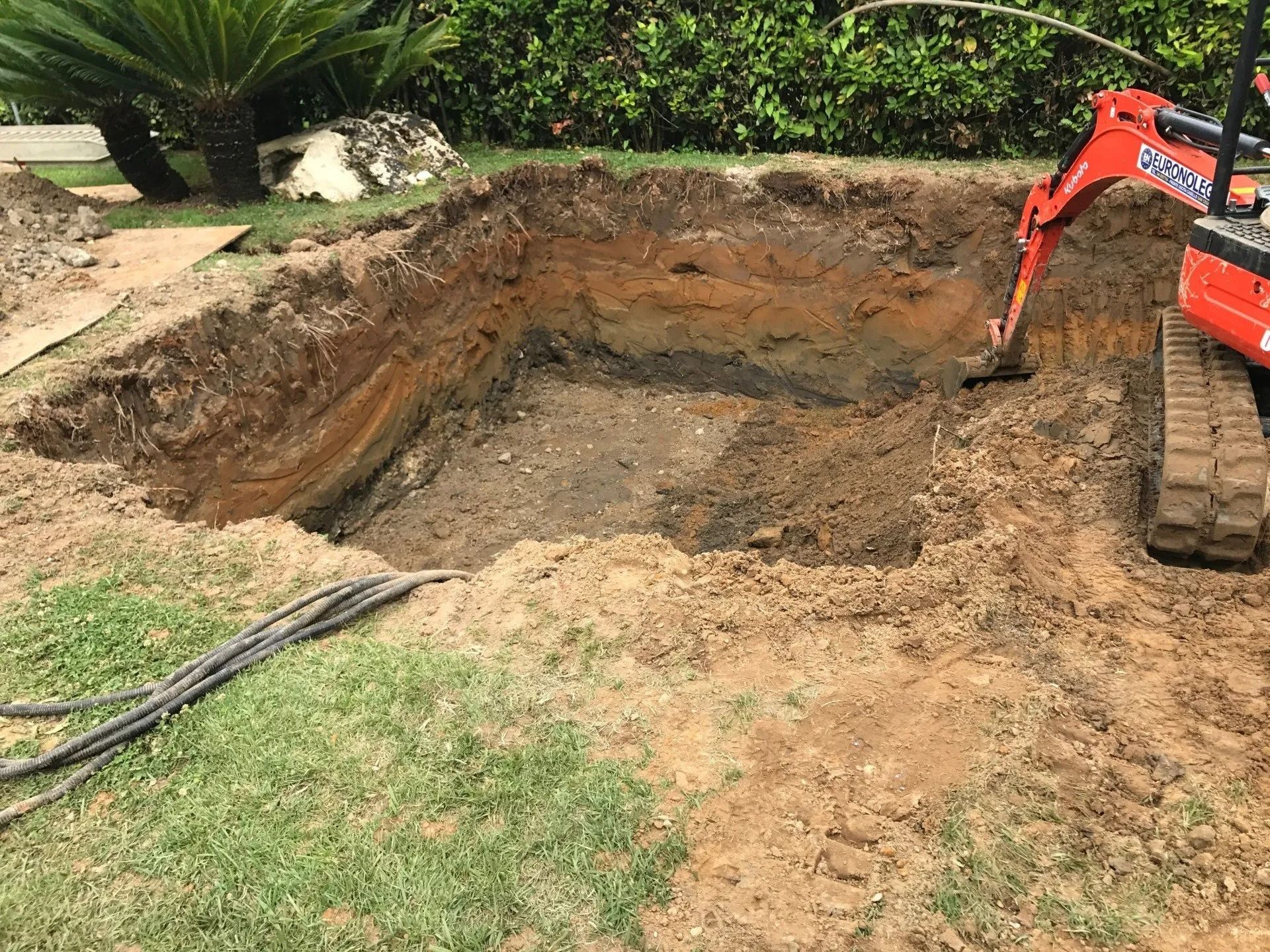 Excavator digging a large rectangular pit in a yard with dirt piles and grass around it