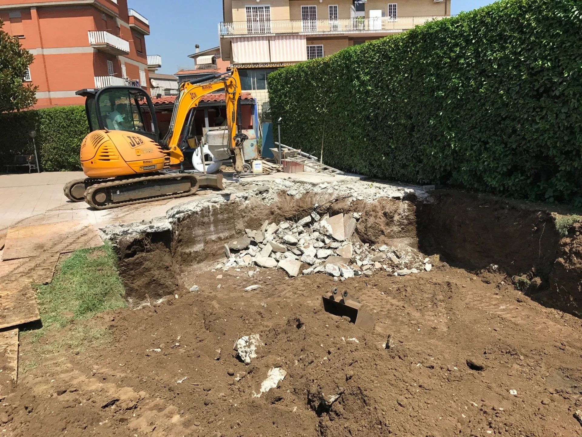 Excavator digging a trench beside a hedge in a residential construction area