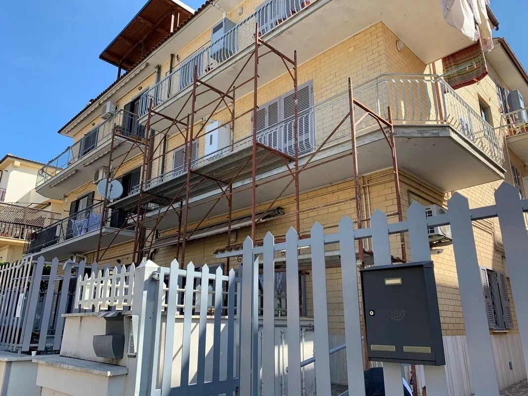 Multi-story apartment building with metal balconies behind a white picket fence under a blue sky