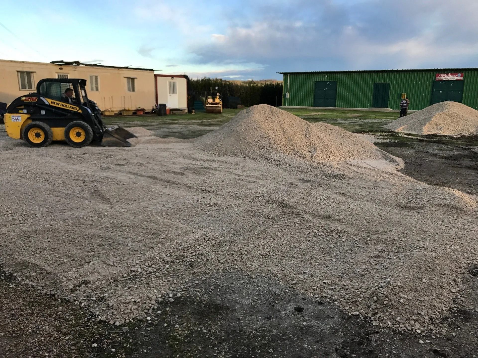 Construction yard with gravel piles, a skid steer, and green site trailers in the background