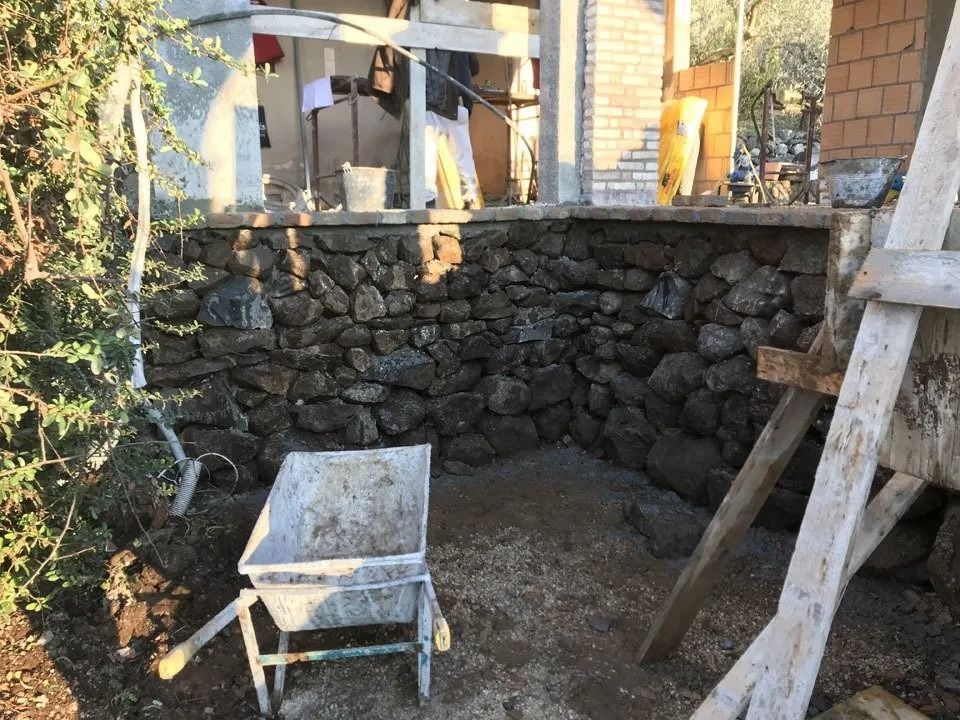 Construction site with a stone retaining wall, wheelbarrow, ladder, and dirt ground outdoors