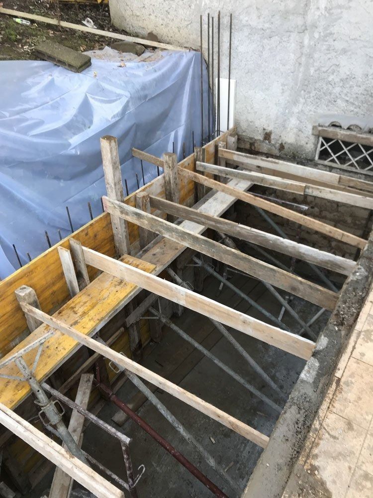 Wooden beams spanning a concrete pit, with a blue tarp-covered stack beside a wall.