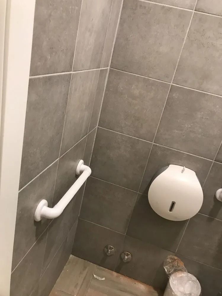 Public restroom stall with gray tiled walls, grab bar, toilet paper dispenser, and trash bin underneath.