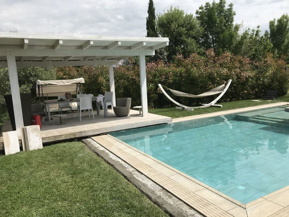 Backyard patio with a pool, white pergola seating area, and hammock beside lush greenery