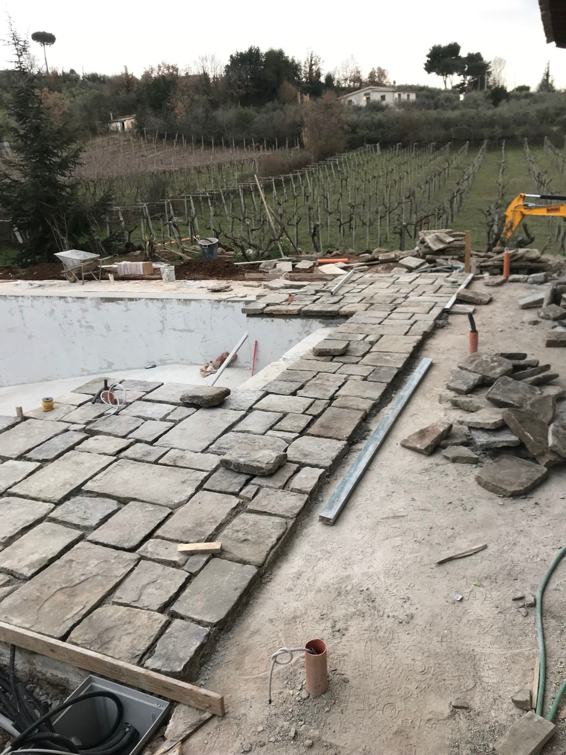 Partially paved construction site beside an empty pool, with stacked stones and an excavator in the background