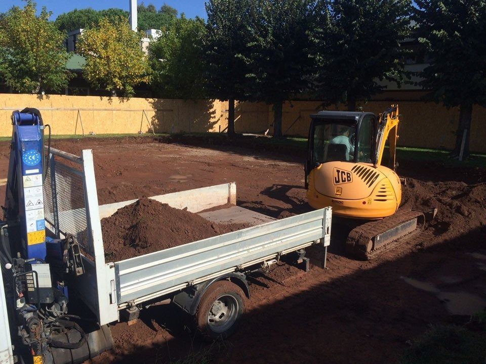 Small excavator beside a white dump trailer in a dirt yard, with a fence and trees in the background.