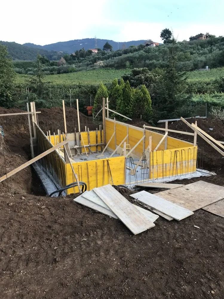 Concrete foundation under construction in a rural hillside with wooden formwork and rebar, surrounded by green fields.