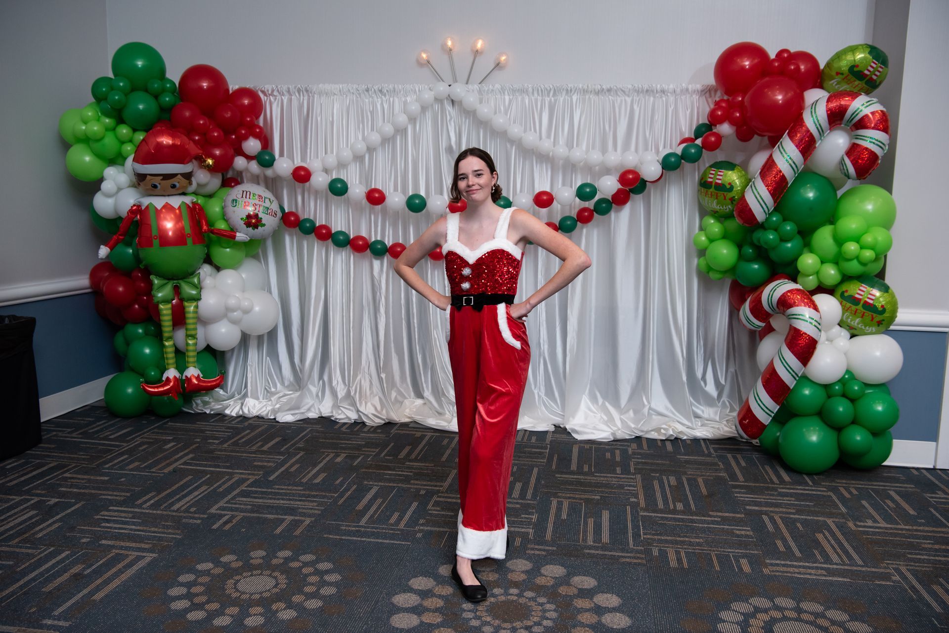 Woman in Santa dress poses in front of Christmas decorations, balloons, and draped fabric.
