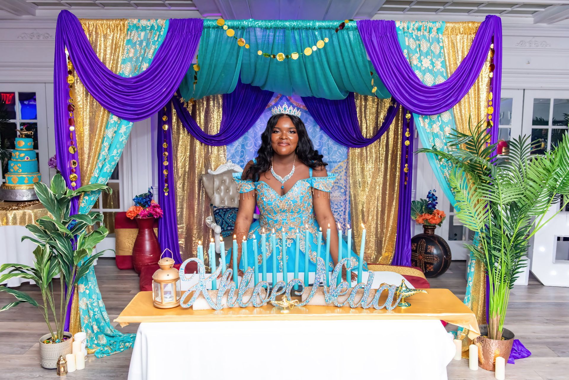 Woman in a turquoise dress with a crown, standing behind a table with candles, in front of a colorful Arabian Nights-themed backdrop.