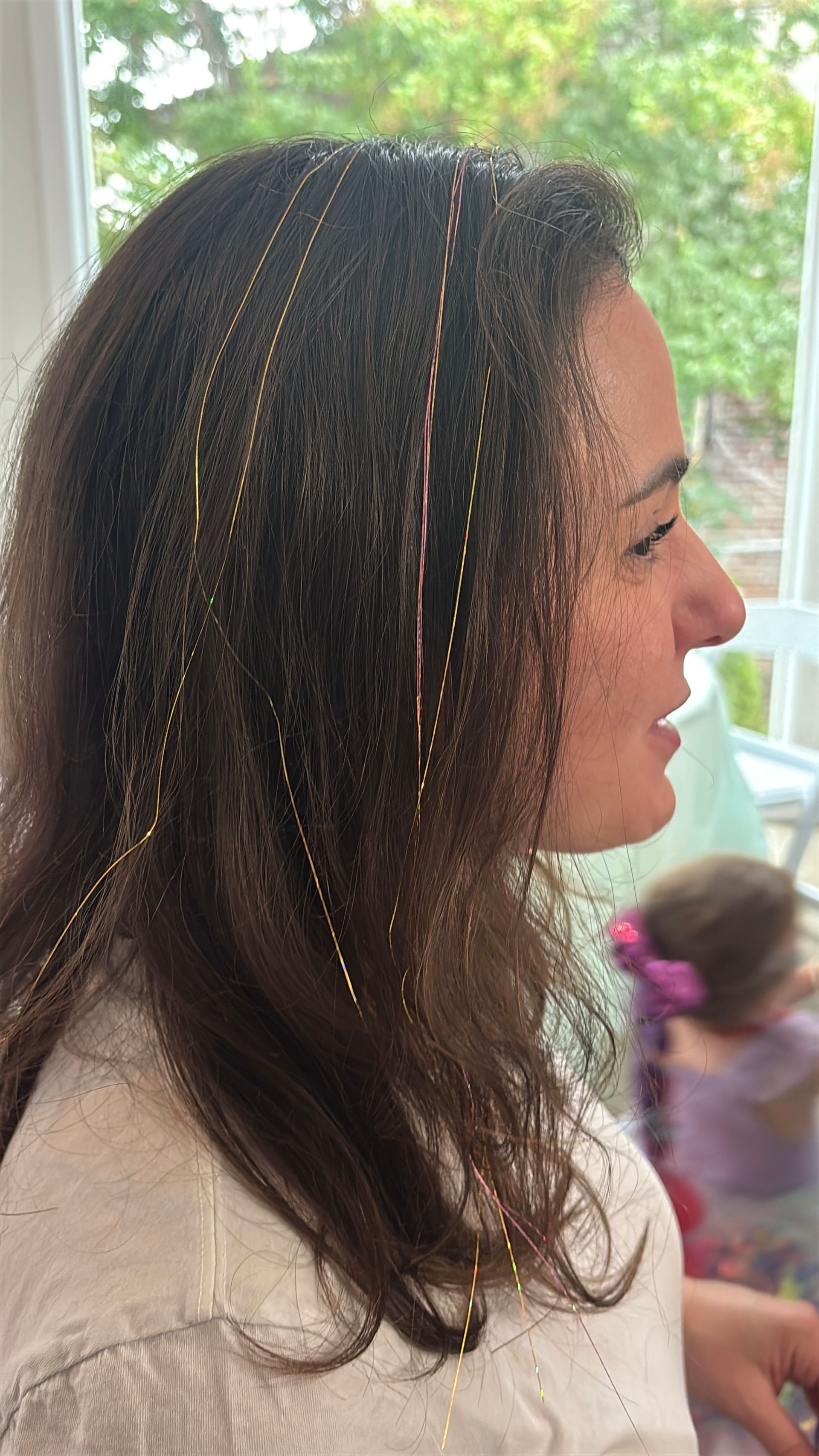 Woman with brown hair, adorned with sparkly strands, smiles near a window.