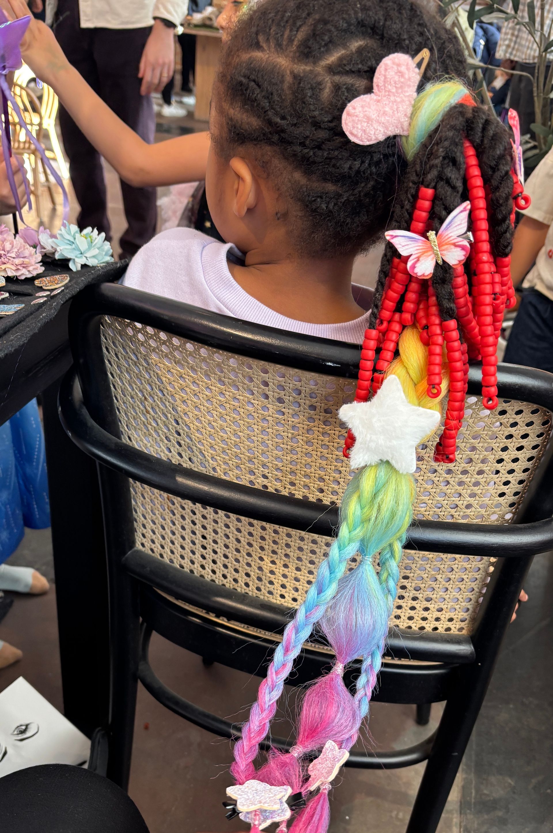 Girl with braided hair decorated with colorful beads, a star, and butterflies, sitting in a chair.