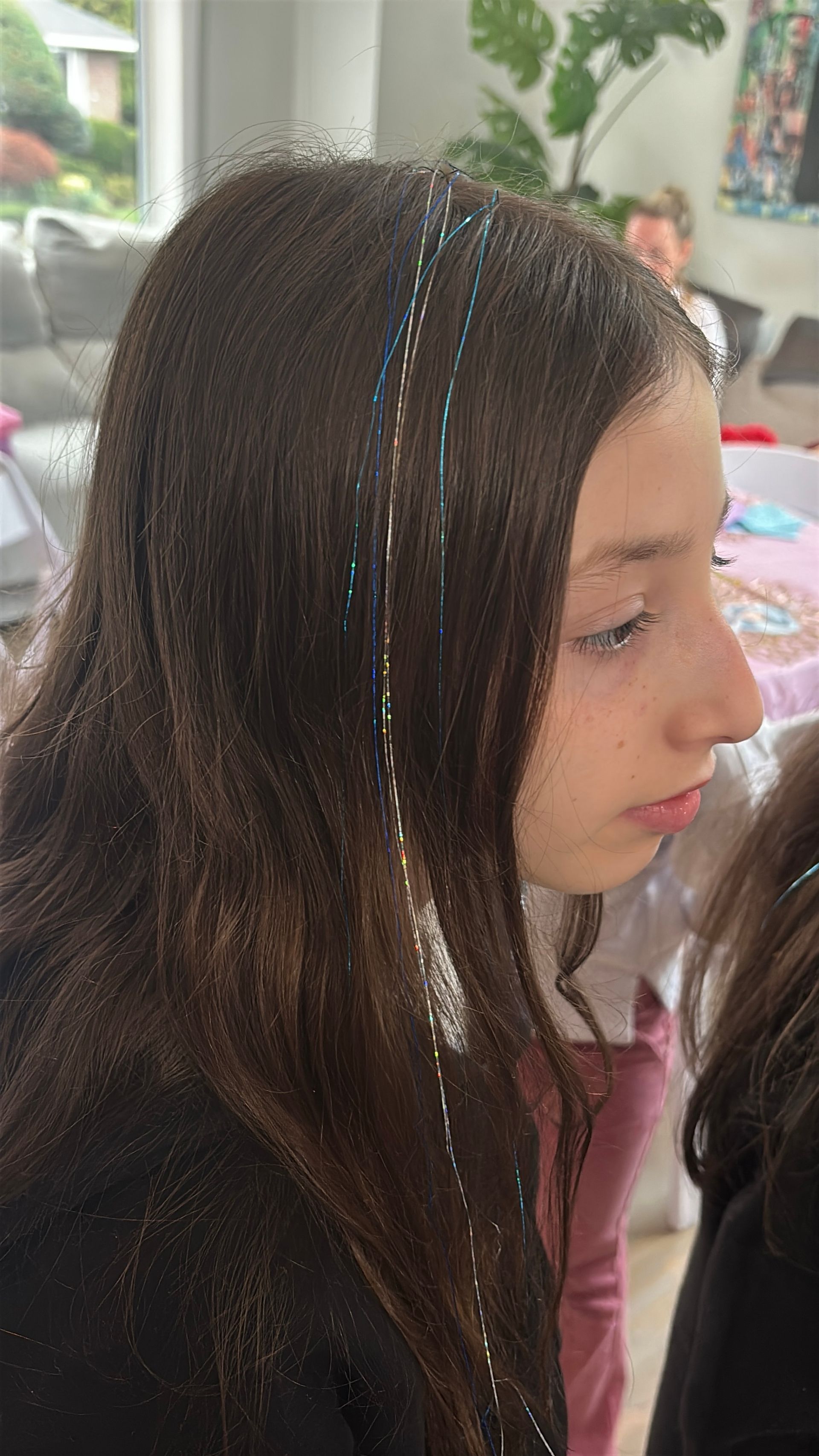 Girl with long brown hair, some strands with glitter, looking to the side. Indoors, blurred background.
