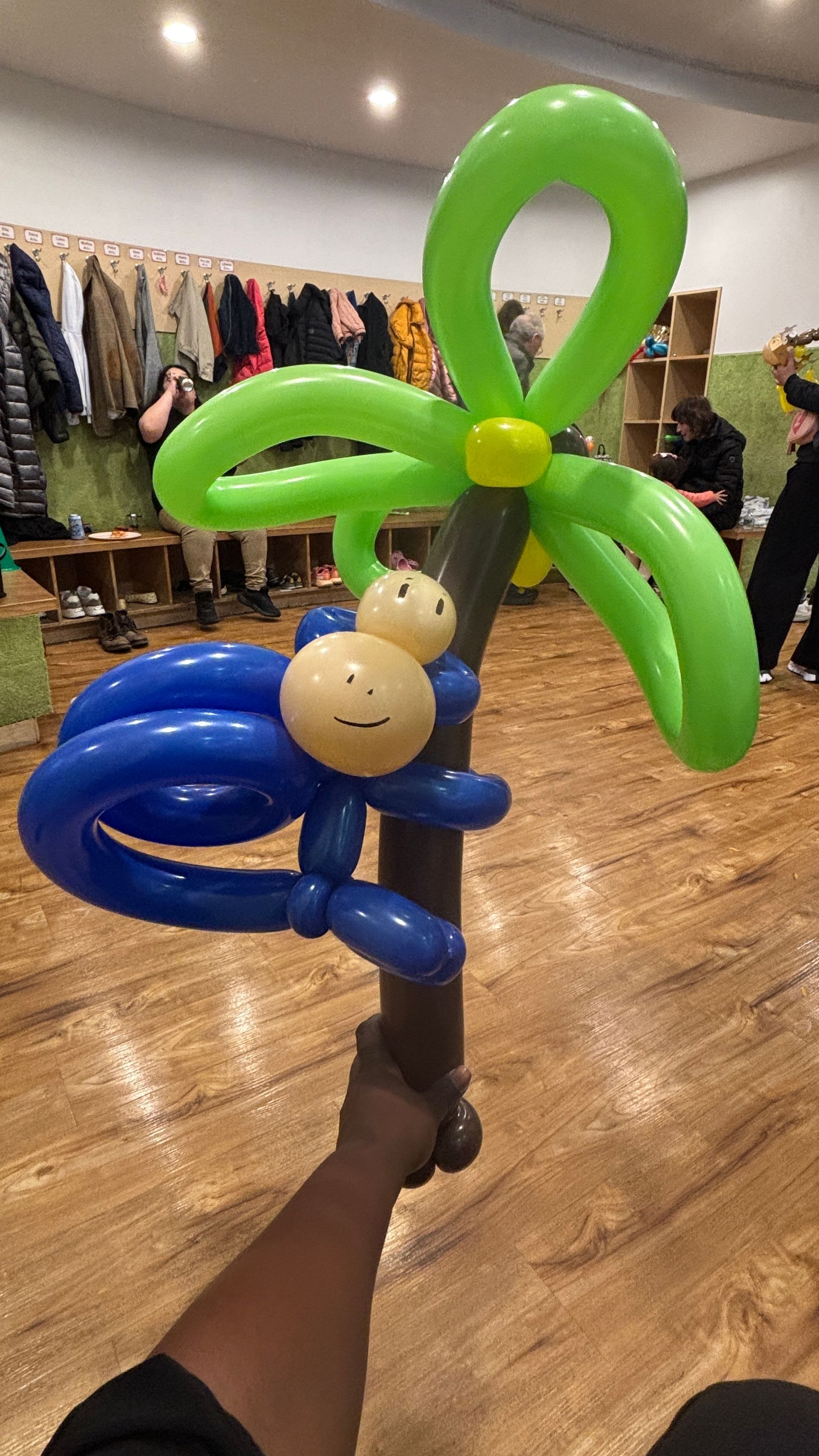 Balloon sculpture of a blue monkey climbing a brown tree with green leaves, held by a hand.