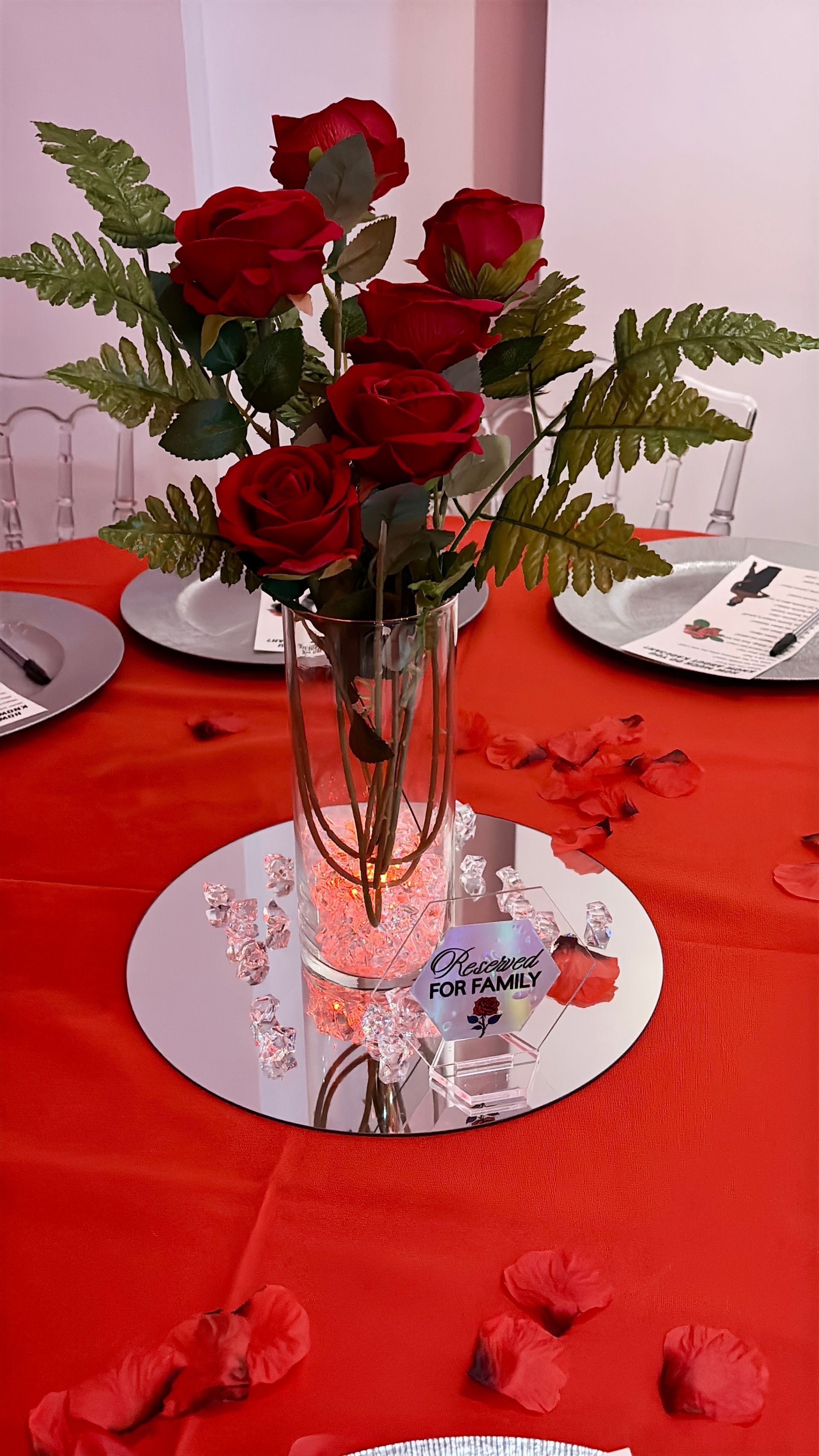 Red roses in a clear vase on a mirrored surface, red tablecloth, place settings.