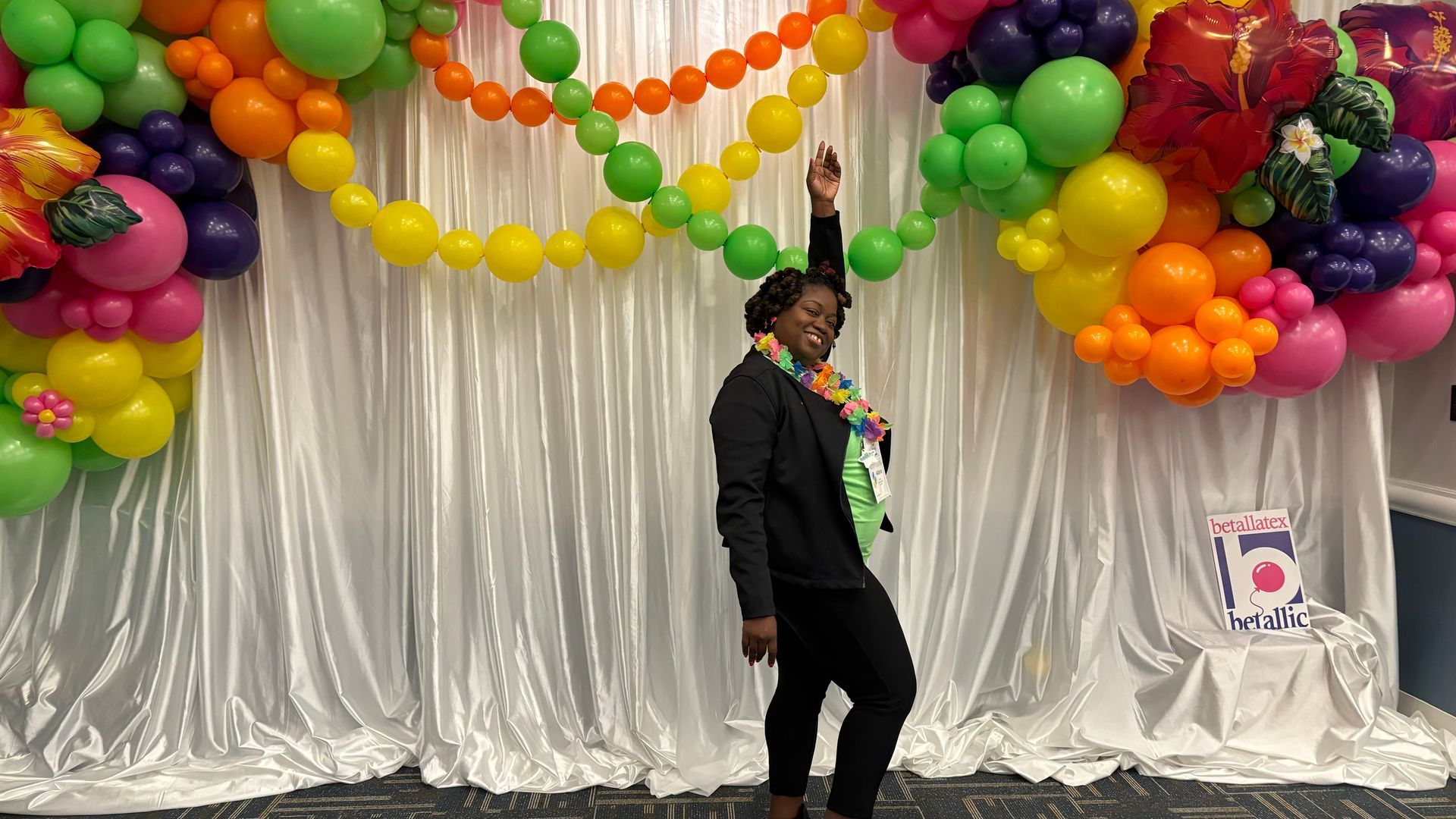Woman with arm raised in front of a white curtain and colorful balloon arch.