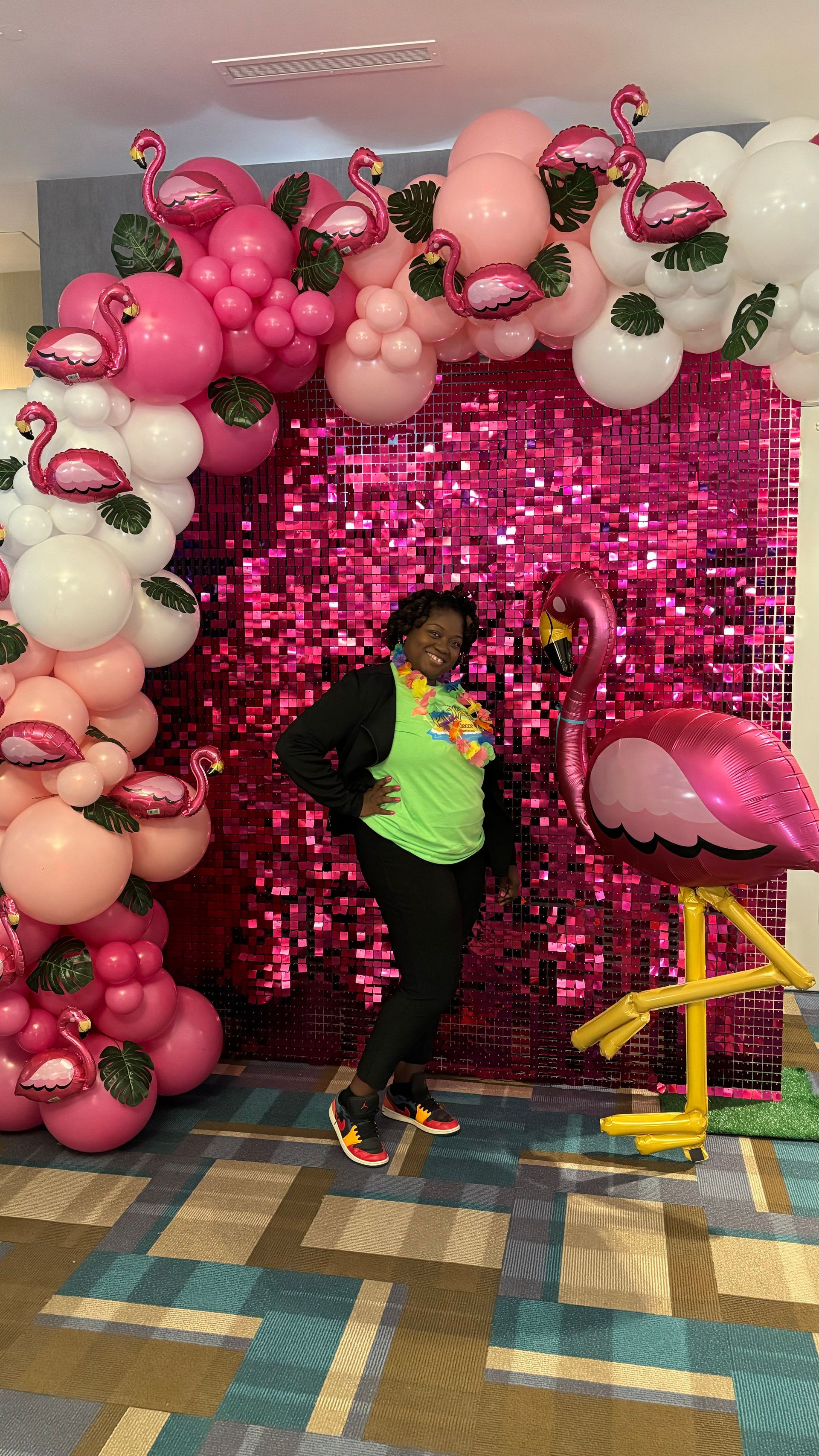 Person posing next to a flamingo backdrop, wearing a lei. Pink sequin wall with balloon arch and inflatable flamingo.