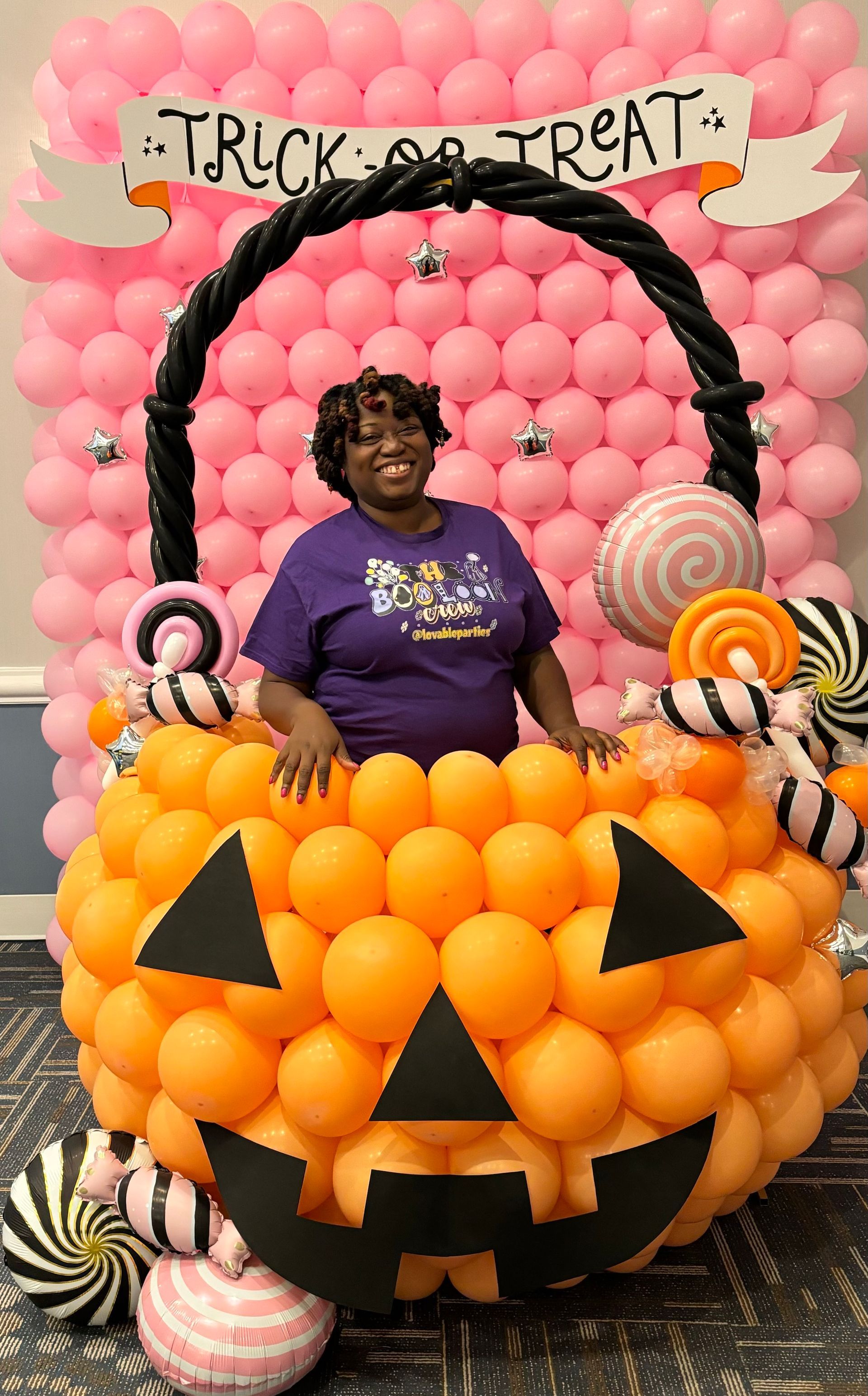 Woman in purple shirt in a balloon pumpkin basket, in front of a pink balloon wall, with a 