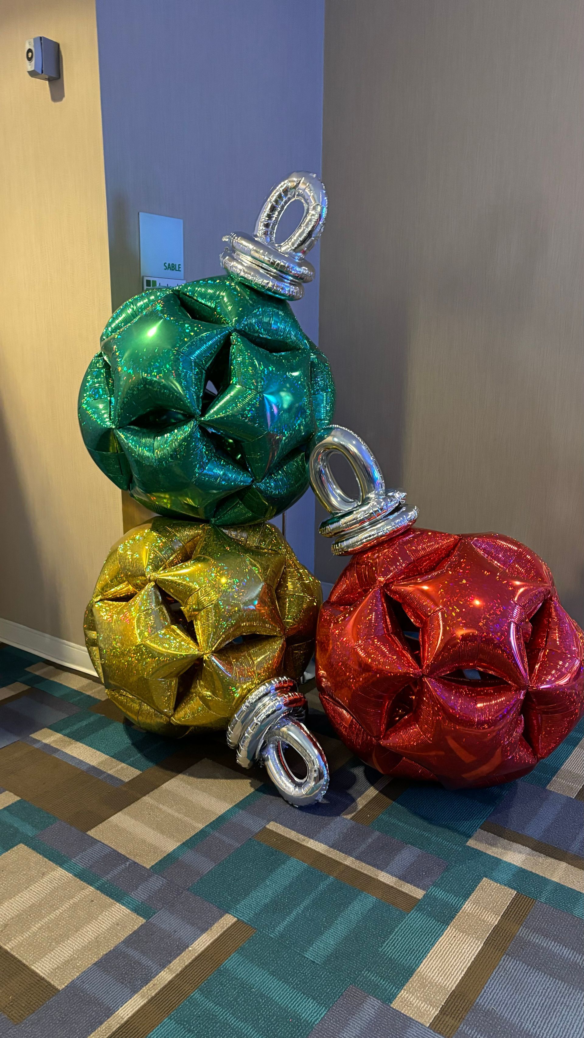 Three large, colorful Christmas ornament balloons stacked on a patterned floor.