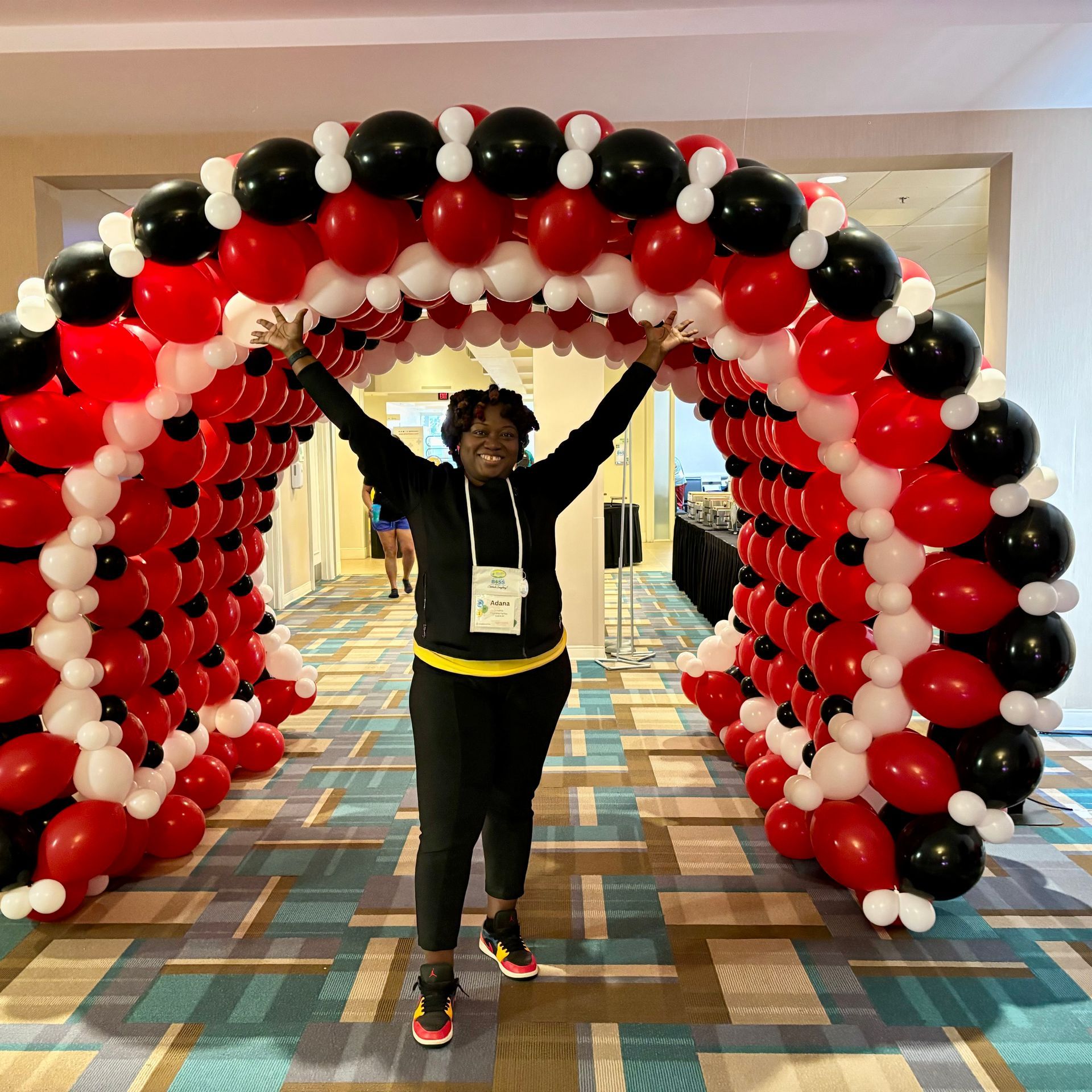 Woman raises arms, standing inside red, black, and white balloon archway in a hallway.