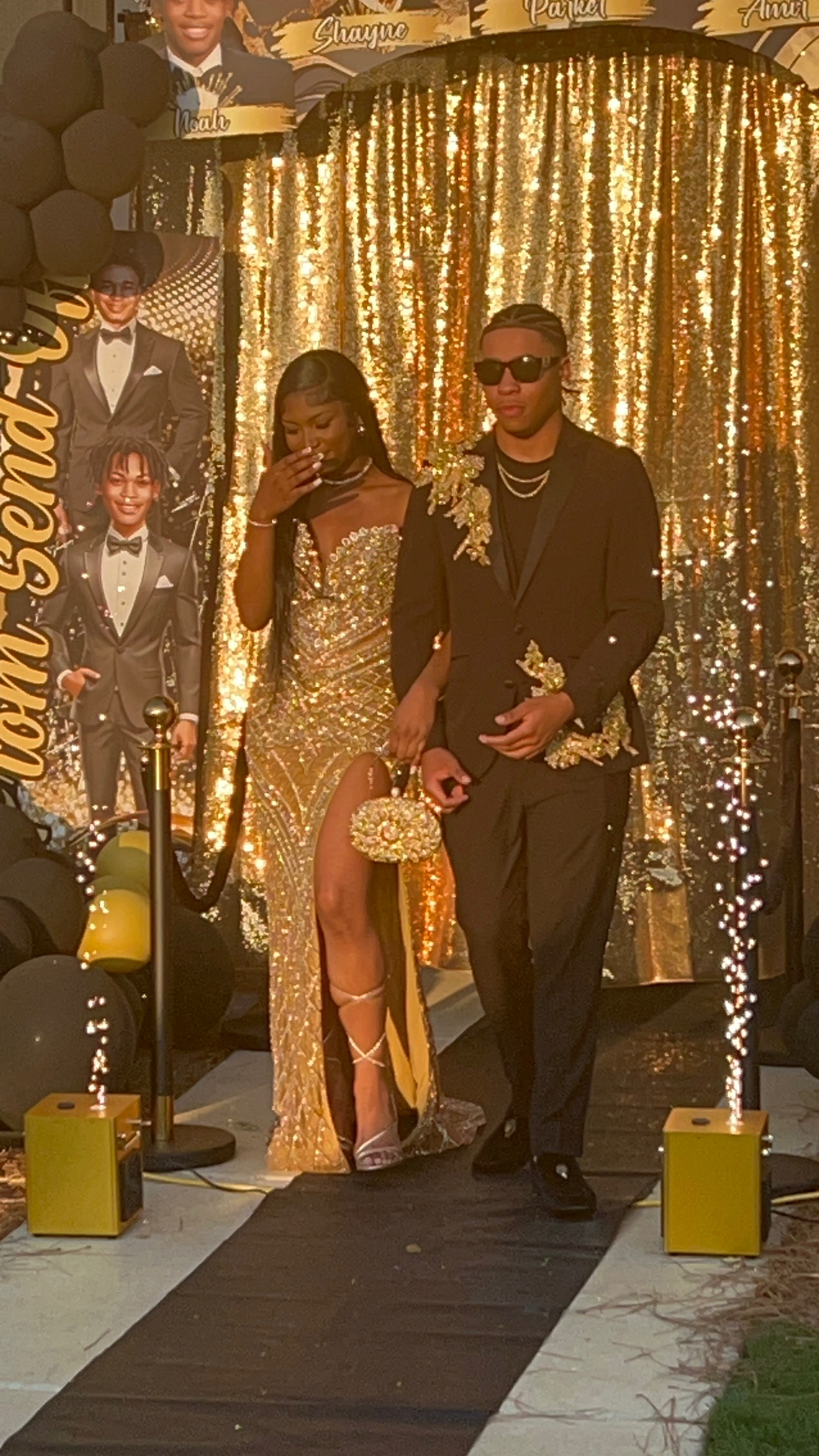 Couple in formal wear walk down a red carpet at a golden-themed event, sparks fly.