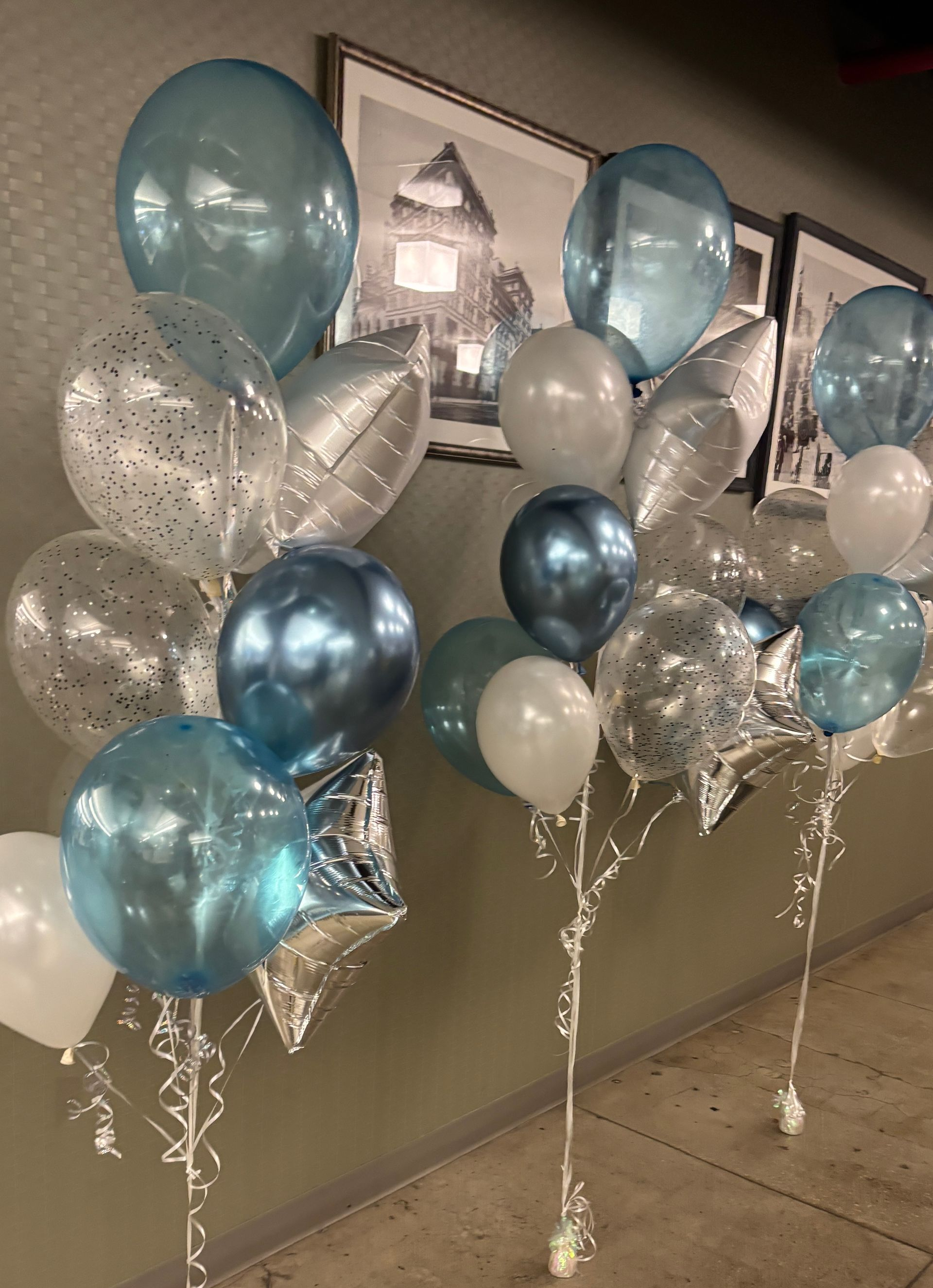 Two balloon bouquets: light blue, silver, and white balloons, some star-shaped, against a wall.