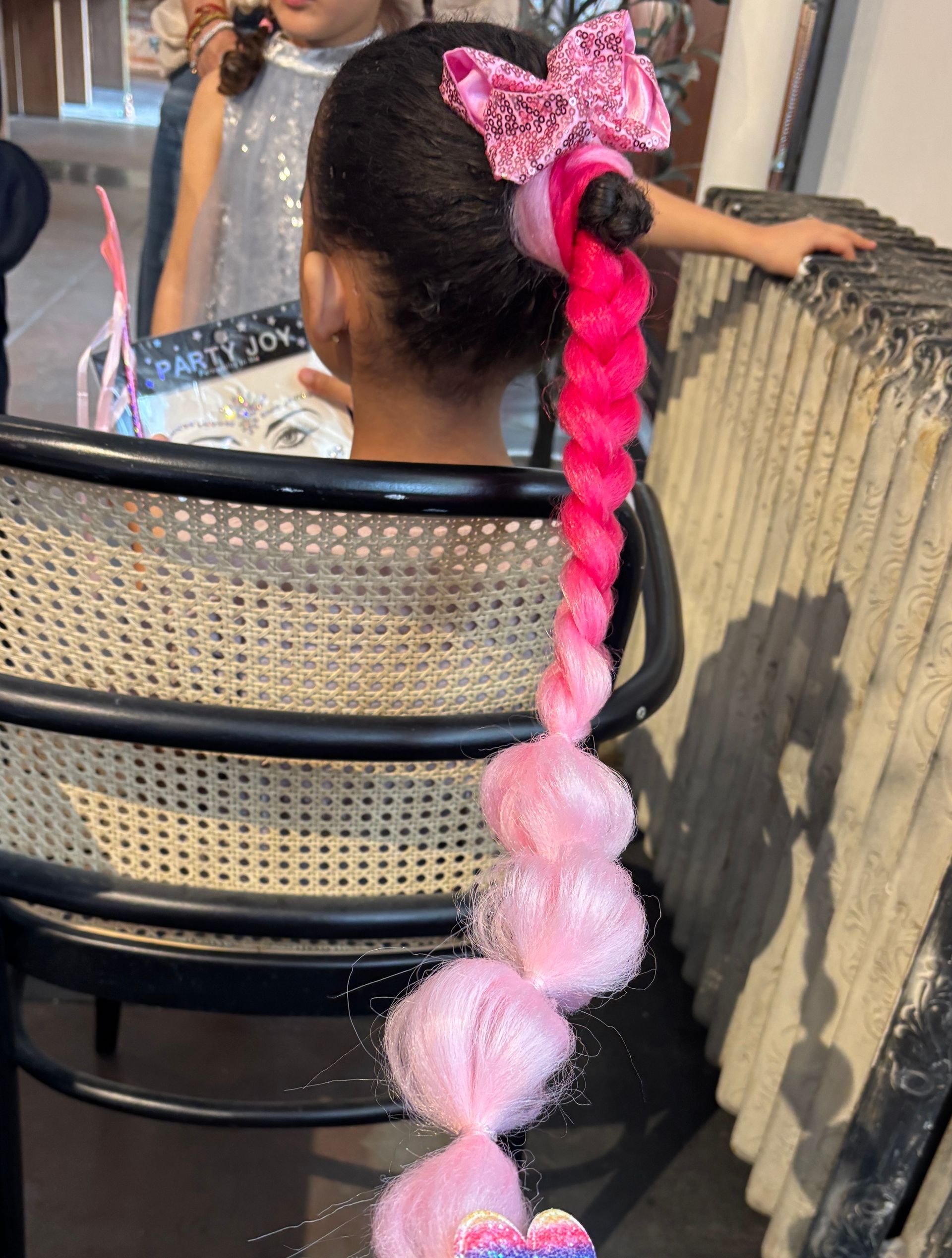 Girl with a pink and magenta braided hairstyle with a butterfly bow, sitting in a chair.