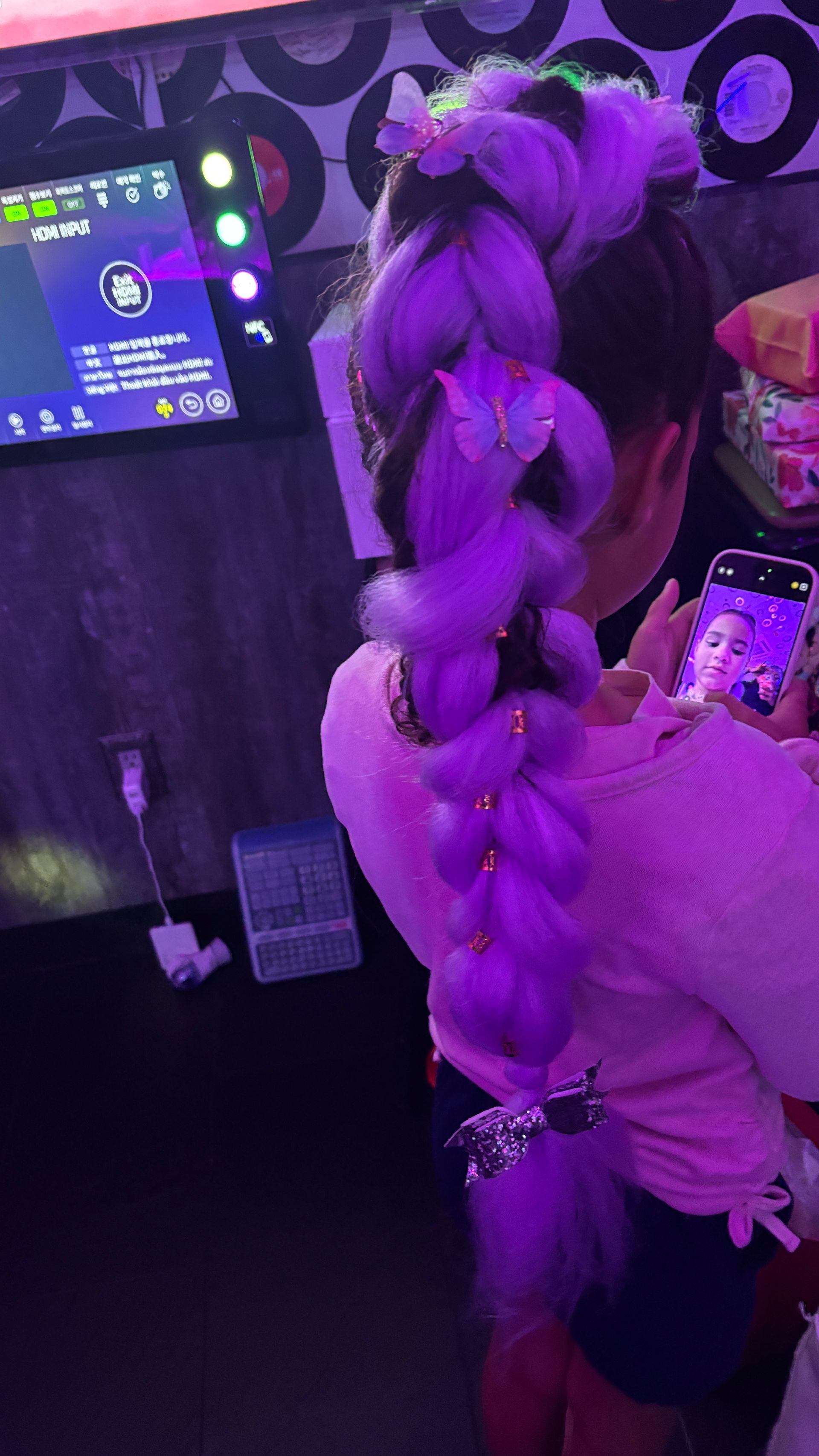Young person with long braided purple hair, adorned with butterfly clips and beads, in a purple-lit setting.