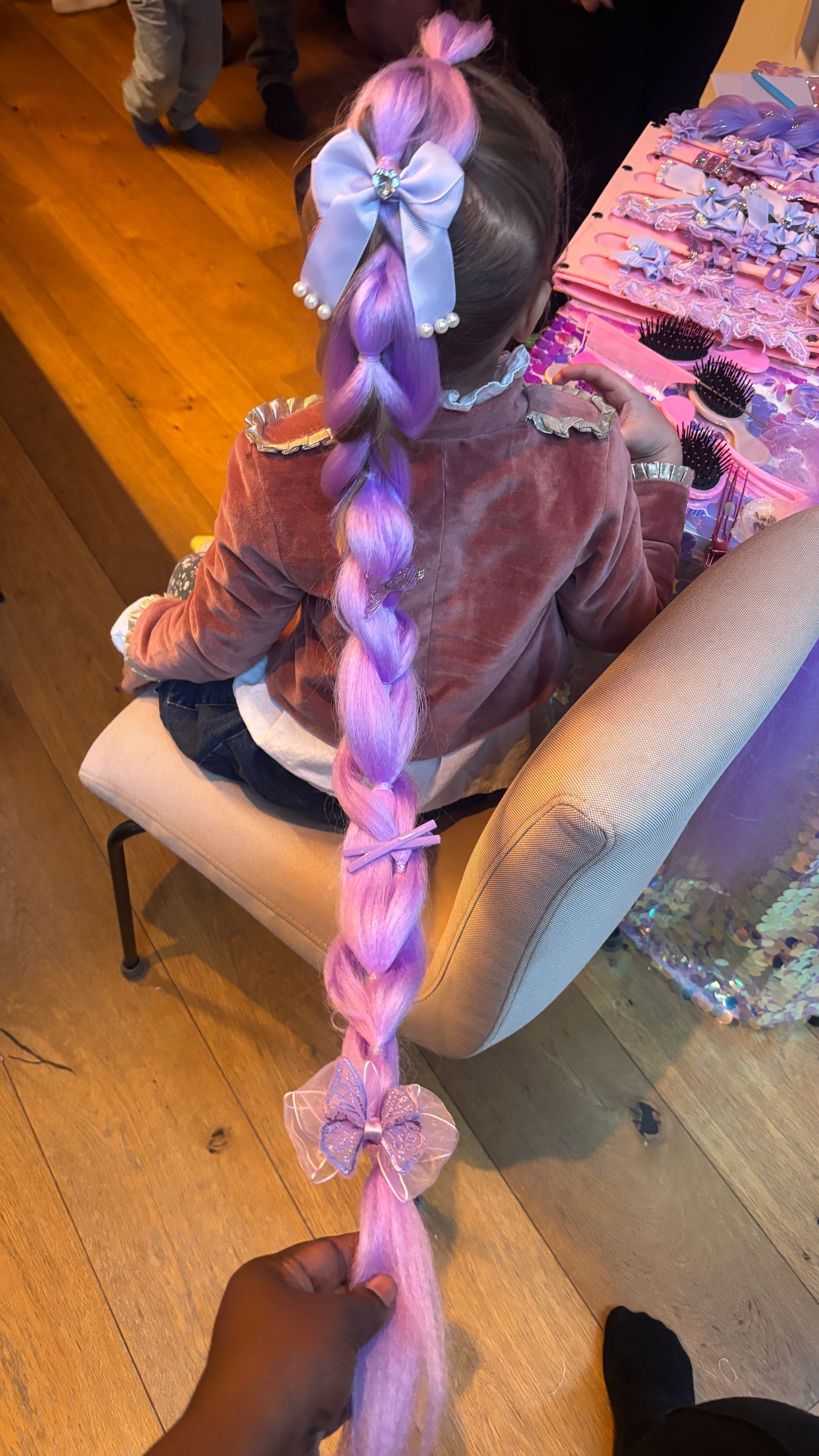Child with long purple braid and bows, seated. A hand holds the braid's end.