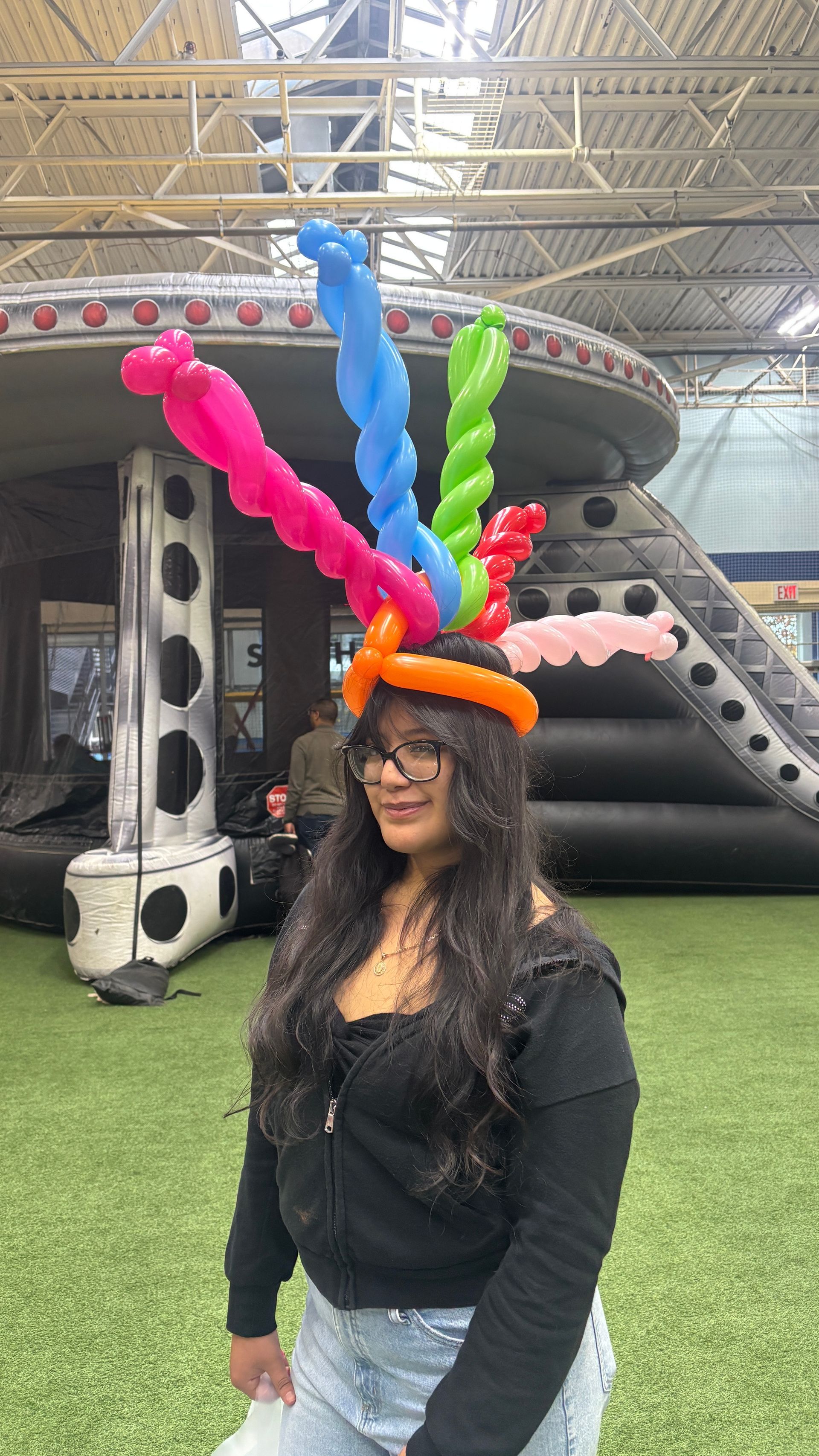 Woman smiling, wearing a colorful balloon hat, standing in front of an inflatable structure and green turf.