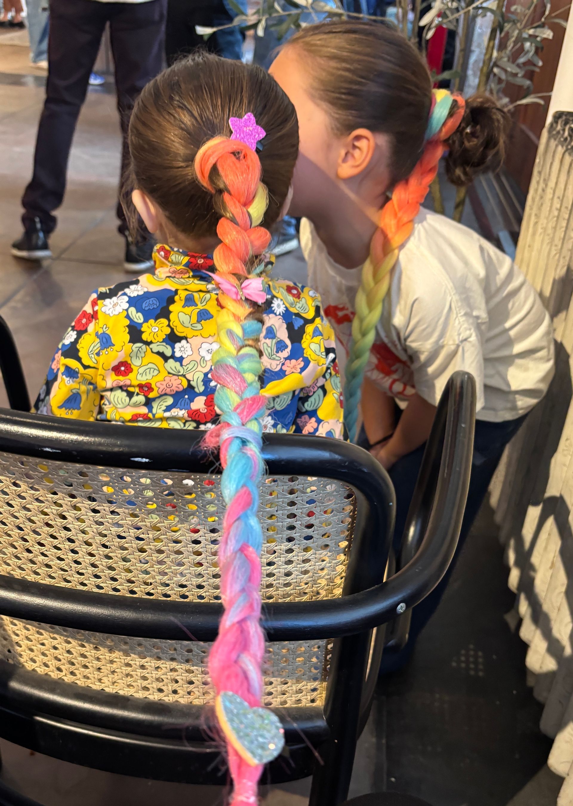 Two children with rainbow braids, one whispering into the other's ear.