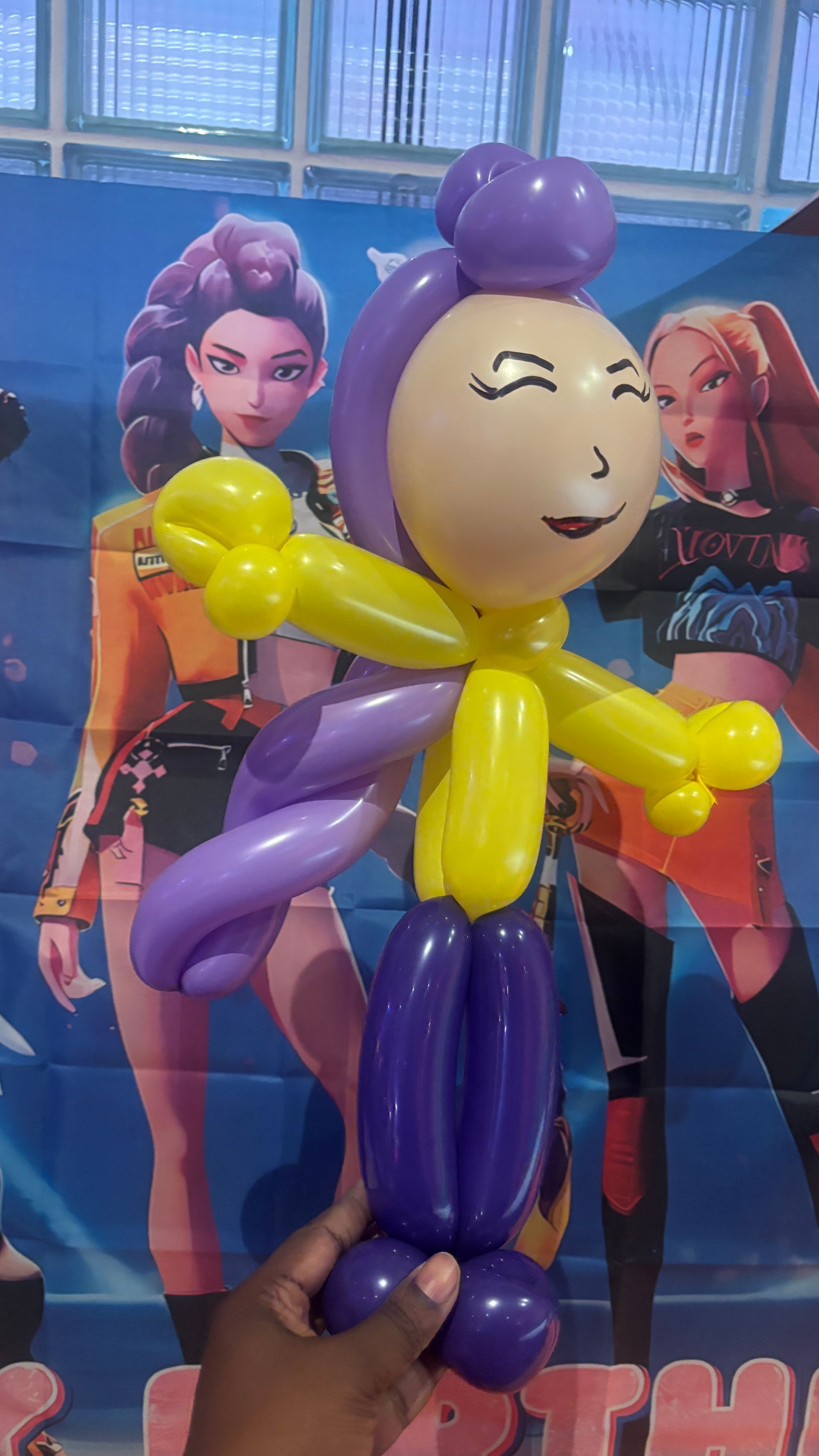 Balloon sculpture of a smiling figure with purple and yellow limbs and hair, held in front of a backdrop with two women.