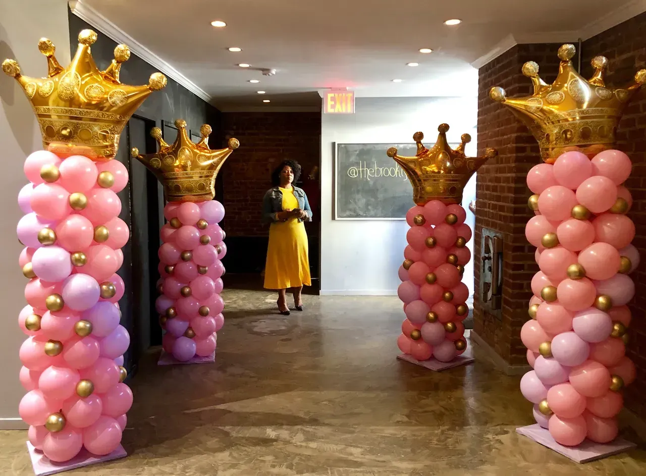 A woman in a yellow dress stands in a hallway surrounded by balloons and crowns