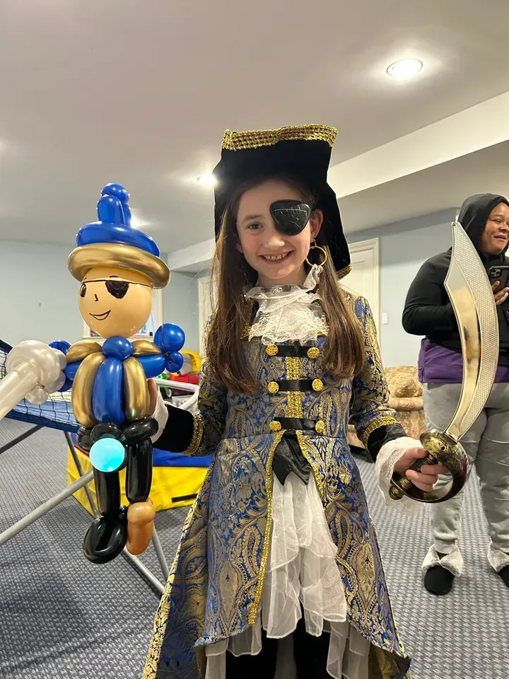 Girl in pirate costume holding a balloon figure and toy sword, smiling indoors.
