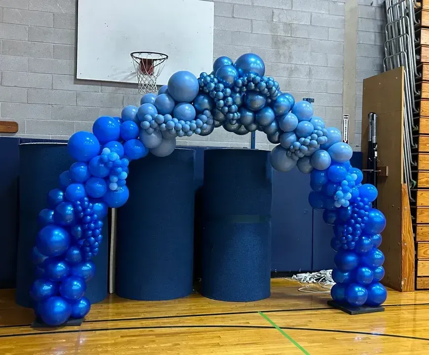 A blue and silver balloon arch in a gym with a basketball hoop in the background.